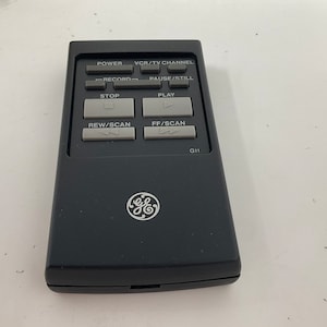 May include: A black GE VCR/TV remote control with gray buttons. The remote has buttons for power, record, play, stop, rewind/scan, and fast forward/scan. The GE logo is at the bottom.