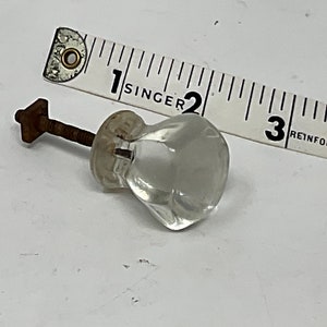 May include: Clear glass cabinet knob with a metal screw and a rusty, square-shaped nut. A white measuring tape with black markings and the word "SINGER" is next to the knob. The knob is approximately 1 inch in diameter.