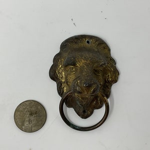 May include: Antique brass lion head door knocker with a circular ring. The lion's face is detailed with a textured mane and facial features. A coin is next to the knocker for size comparison.