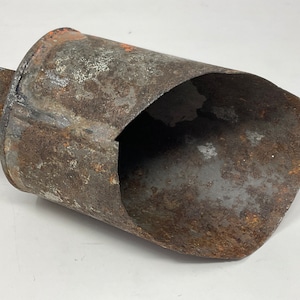 May include: A weathered, rusty metal scoop with a cylindrical body and a short, rectangular handle. The scoop has a large opening on one side, revealing the interior. The metal shows signs of age and wear, with areas of rust and discoloration.