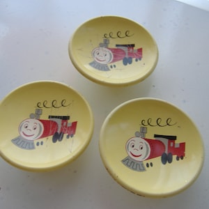 May include: Three small, yellow, vintage plates with a cartoon train design. Each plate features a smiling red train with black and gray accents. The plates are slightly chipped, showing signs of age.