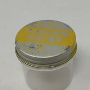 May include: A clear glass jar with a metal lid. The lid is silver with a yellow stripe across the top. The jar is empty and sits on a white surface.