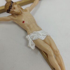 May include: A detailed figurine of Jesus Christ on a wooden cross. The figure is depicted with a crown of thorns, a white loincloth, and visible wounds. The cross is made of light brown wood.