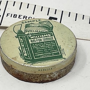 May include: A small, round, antique tin of Williams Anti-Pain Ointment. The tin has a cream-colored lid with green text and graphics, including the product name and a depiction of the ointment. The base is rusty brown. A ruler is in the background.