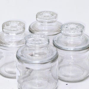 May include: Four clear glass jars with matching lids. The jars are cylindrical with a slightly rounded base. The lids are flat with a decorative, raised pattern in the center. The jars are empty and appear to be clean.