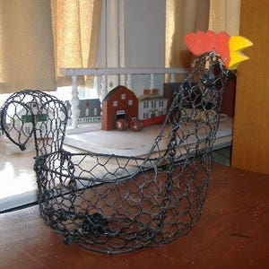 May include: A black wire chicken-shaped basket with a red comb and yellow beak. The basket is made of a wire mesh and sits on a brown surface. The chicken basket is a decorative item.