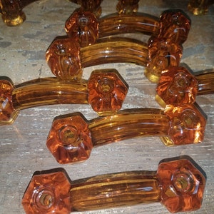 May include: A collection of amber-colored glass drawer pulls, each featuring a curved handle design with hexagonal mounting ends. The glass is translucent and smooth, showcasing a warm, golden-brown hue throughout the hardware pieces.