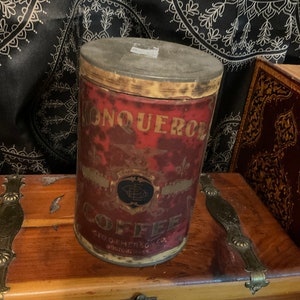 May include: Vintage metal coffee can with a red label that says "Conqueror Coffee" and a gold seal with a globe design. The can is slightly rusted and has a worn, aged look.