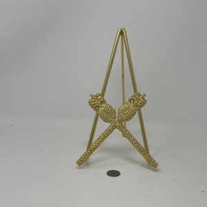 May include: A gold metal display stand with a triangular base and two decorative pineapple-shaped arms.