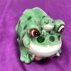 May include: A green ceramic frog figurine with brown eyes and white spots. A smaller green frog sits on the back of the larger frog.