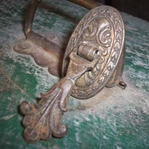 May include: An antique brass drawer pull with an ornate oval backplate featuring a floral design. The pull has a decorative, leaf-like handle and a curved support. The metal shows signs of age and wear, with a patina finish.