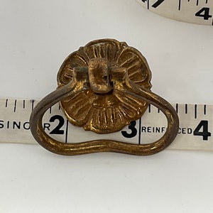 May include: A vintage brass drawer pull with a floral design and a curved handle. The pull is approximately 3 inches wide and is set against a white background with a measuring tape.