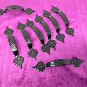 May include: A collection of black, hammered-finish metal handles. Each handle features a curved bar with decorative heart-shaped ends. The handles are arranged on a purple surface, showcasing their design and texture.