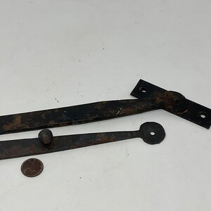 May include: Two antique, black metal hinges with a rustic, weathered appearance. One hinge is a long, straight bar with a circular end, and the other has a rectangular plate with screw holes. A penny is next to the hinges.
