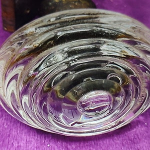 May include: Clear glass bottle stopper with a spiral design. The stopper has a rounded shape and is partially transparent, revealing a dark interior. The glass has a textured surface with concentric circular patterns.