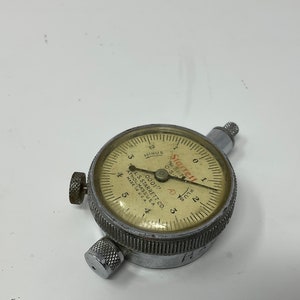 May include: A vintage Starrett dial indicator gauge with a silver metal casing and a cream-colored dial. The dial features black numbers and markings, with the text "Starrett" and "Athol, Mass. U.S.A." visible. The gauge has three adjustment knobs.