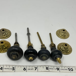 May include: Four antique black wooden knobs with brass accents and matching decorative brass backplates. The knobs have varying stem designs. A ruler with inch measurements is in the foreground.