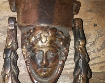 May include: A vintage brass door knocker with a detailed design of a face with a crown and a leafy wreath. The knocker is attached to a decorative scrollwork design.