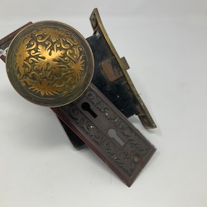 May include: Antique brass door knob with an ornate floral design. Includes a matching keyhole plate and a black and gold door latch. The hardware is likely from an older home.