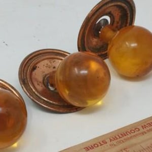 May include: Three vintage amber glass door knobs with aged copper-colored backplates. The knobs are round and have a warm, translucent appearance. A wooden ruler with printed text is in the foreground. The knobs are likely for antique furniture.