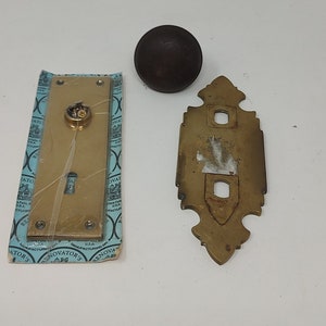 May include: An assortment of vintage brass door hardware, including a doorknob, a keyhole plate, and a decorative backplate. The keyhole plate is still in its original packaging. The hardware is likely from the early to mid-20th century.