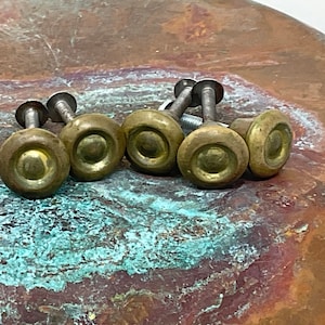 May include: A collection of five antique brass knobs with silver screws. The knobs have a rounded, circular design with a central indentation. The brass has a warm, aged patina, suggesting vintage hardware.
