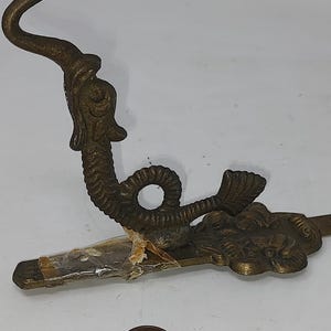 Puede incluir: Un gancho de pared antiguo de tono latón con un diseño detallado de dragón o serpiente marina. El gancho tiene una parte superior curvada y una base decorativa con elementos florales. El gancho está unido a una barra plana y está envuelto en plástico transparente.