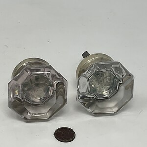 May include: Two clear, faceted glass door knobs with silver-toned metal bases. The knobs have an octagonal shape and are displayed on a white background. A small, brown coin is at the bottom.