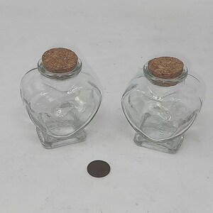 May include: Two clear glass jars shaped like hearts, each topped with a round cork stopper. The jars have a wide, rounded upper section that tapers down to a square base. A small, dark brown circular coin is positioned between the two glass jars.