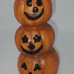 May include: A stacked Halloween decoration featuring three orange pumpkins with carved faces. Each pumpkin has a smiling face with cut-out eyes and a mouth. The top pumpkin has a brown stem.