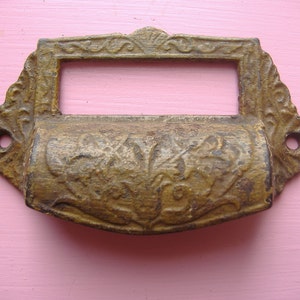 May include: An antique brass drawer pull with an ornate, scrollwork design. The rectangular pull has a central opening for a label and two holes for mounting. The brass has a weathered, aged appearance, with visible wear and tear.