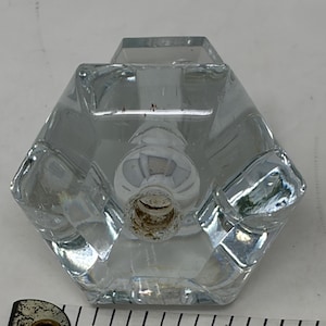 May include: A clear, faceted glass knob with a hexagonal shape and a central metal-lined opening. The knob is positioned above a white measuring tape that shows the object is approximately 2 inches wide at its base, featuring the text SINGER.