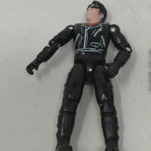 May include: A plastic action figure with dark hair and a black mask, wearing a black leather-style jacket with white zipper details over a red shirt, paired with black pants and boots. The figure has articulated joints at the shoulders and elbows.