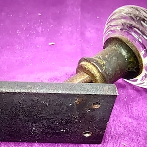 May include: Antique glass doorknob with a clear, swirled design. The knob is attached to a dark, rectangular metal plate with screw holes. The metal has a weathered, aged appearance.