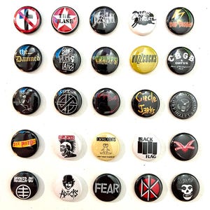 Puede incluir: Una colección de veinticinco botones redondos y coloridos con logotipos de bandas e imágenes de punk rock. Los botones muestran texto y gráficos en varios colores, incluyendo rojo, negro, blanco y amarillo. Los botones están dispuestos en un patrón de cuadrícula.