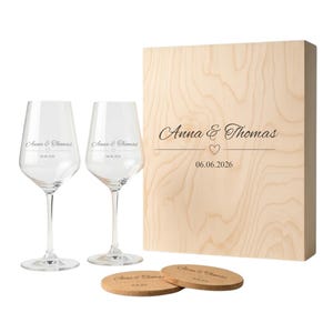 Könnte beinhalten: Eine Holzgeschenkbox, zwei Weingläser und zwei Untersetzer. Die Box ist mit "Anna & Thomas", einem Herz und "06.06.2026" graviert. Die Gläser und Untersetzer tragen ebenfalls die Namen und das Datum. Die Gläser sind klar.