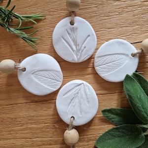 May include: Four white clay ornaments with different leaf and stem imprints, each hanging from a twine string with a wooden bead. The ornaments are on a wooden surface with sprigs of rosemary and sage.