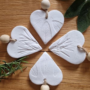 May include: Four white clay heart ornaments with a leaf design. Each heart is strung with twine and a wooden bead. The hearts are arranged in a circle on a wooden surface.
