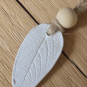 Könnte beinhalten: Ein weißes, blattförmiges Ornament mit detaillierten Adern, befestigt an einer Schnur aus Naturfaser und einer Holzperle. Das Ornament befindet sich vor einem hellbraunen Holzgrund.