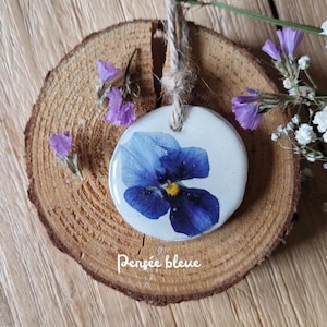 May include: A circular, off-white ceramic pendant featuring a pressed blue pansy flower and a jute string. The pendant is displayed on a wooden slice, with small purple flowers and the text "Pensée bleue".