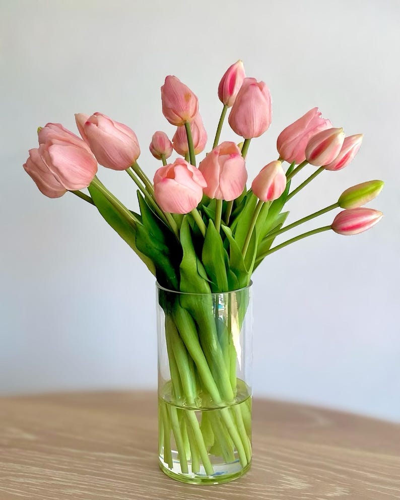 K&ouml;nnte beinhalten: Ein Strau&szlig; hellrosa Tulpen mit gr&uuml;nen Stielen und Bl&auml;ttern in einer klaren, zylindrischen Glasvase mit Wasser. Die Tulpen befinden sich in verschiedenen Bl&uuml;hstadien, von geschlossenen Knospen bis hin zu voll ge&ouml;ffneten Bl&uuml;ten mit rosa Bl&auml;ttern.