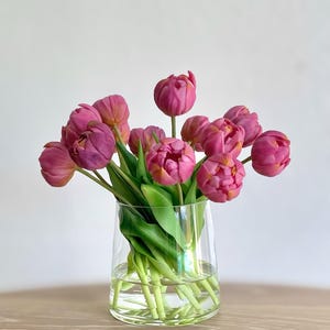 May include: A bouquet of pink tulips in a clear glass vase. The tulips are arranged in a loose, natural style, with some of the flowers facing forward and others facing to the side. The vase is sitting on a light brown wooden surface.