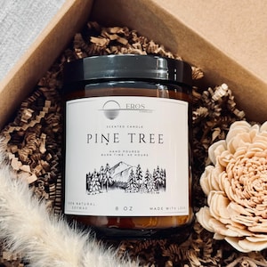 Peut inclure: Une bougie parfumée dans un pot en verre avec un couvercle noir, étiquetée "Pine Tree" avec une illustration de montagne et de forêt. L'étiquette indique également "Hand Poured" et "Burn Time 40 Hours". La bougie est de 227g et faite de cire de soja.