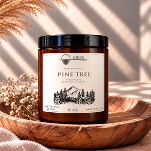 Op de afbeelding: Een kaars in een amberkleurige glazen pot met een zwarte deksel en een wit etiket. Het etiket bevat de tekst EROS CANDLES, PINE TREE, HAND POURED, BURN TIME: 40 HOURS, 100% NATURAL SOYWAX, 8 OZ, MADE WITH LOVE en een bergbosillustratie.