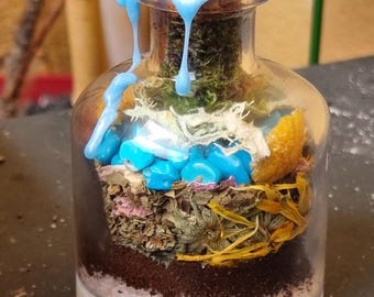 Puede incluir: Una botella de vidrio transparente con un tapón de corcho sellado con cera azul. La botella está llena de capas de hierbas secas, flores y cristales. La botella está sobre una superficie de madera.
