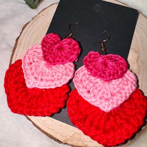 Puede incluir: Un par de pendientes de corazón de ganchillo en rojo, rosa y fucsia. Los pendientes están superpuestos y cuelgan de ganchos dorados.
