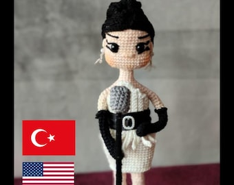 Peut inclure: Patron de poupée amigurumi au crochet d'une chanteuse tenant un microphone. La poupée porte une robe blanche avec une ceinture noire et des gants noirs. Le patron est disponible en format PDF. L'image comprend également le texte "Güneş Handcraft" et des drapeaux de la Turquie et des États-Unis.