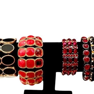 May include: Four bracelets displayed on a stand. The first is black with gold-tone oval links. The second is a wide red faceted stone bracelet with gold-tone settings. The third is a multi-row red crystal bracelet. The fourth is a black crystal bracelet.