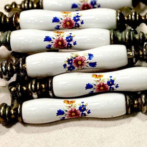 May include: A collection of drawer pulls featuring white ceramic handles with a floral pattern of pink, blue, and yellow flowers. Each handle is flanked by ornate, aged brass-toned metal hardware with a tiered, spindle-like design on both ends.