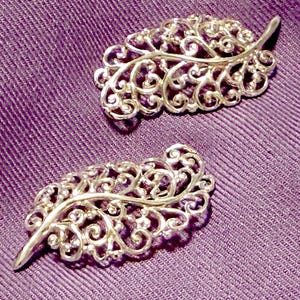 May include: Two silver-toned leaf-shaped brooches with intricate, openwork designs. The brooches are displayed on a textured, purple fabric background. The leaves have detailed, swirling patterns.